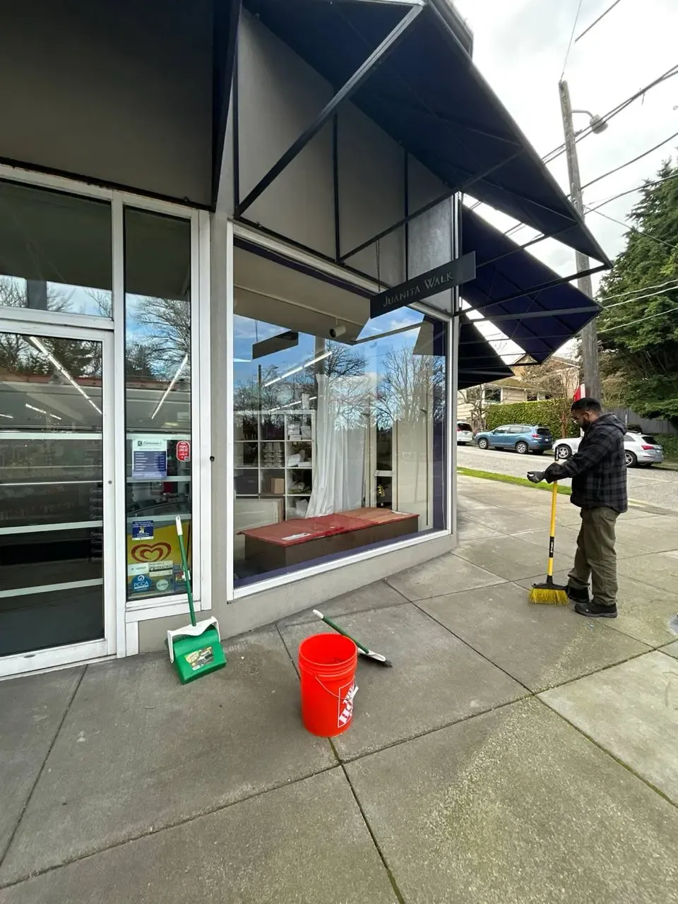 A person with a broom outside a glazed storefront, beside cleaning equipment.