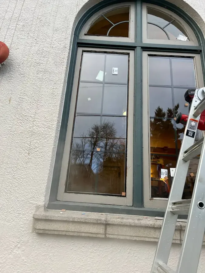 Arched residential window with replacement glass labels and a ladder beside the frame.