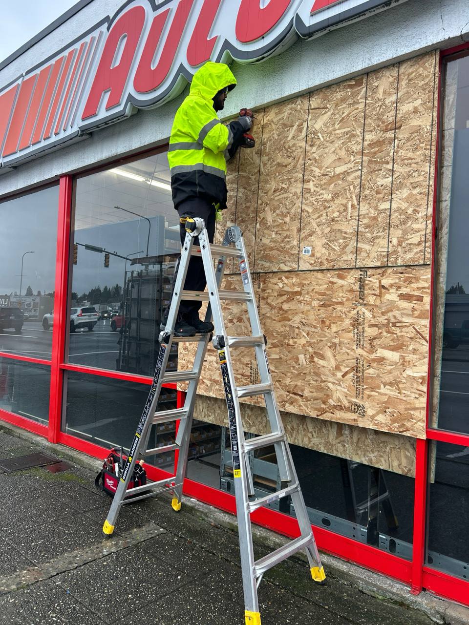 Worker on a ladder beside a timber board covering an opening in a red retail frontage.