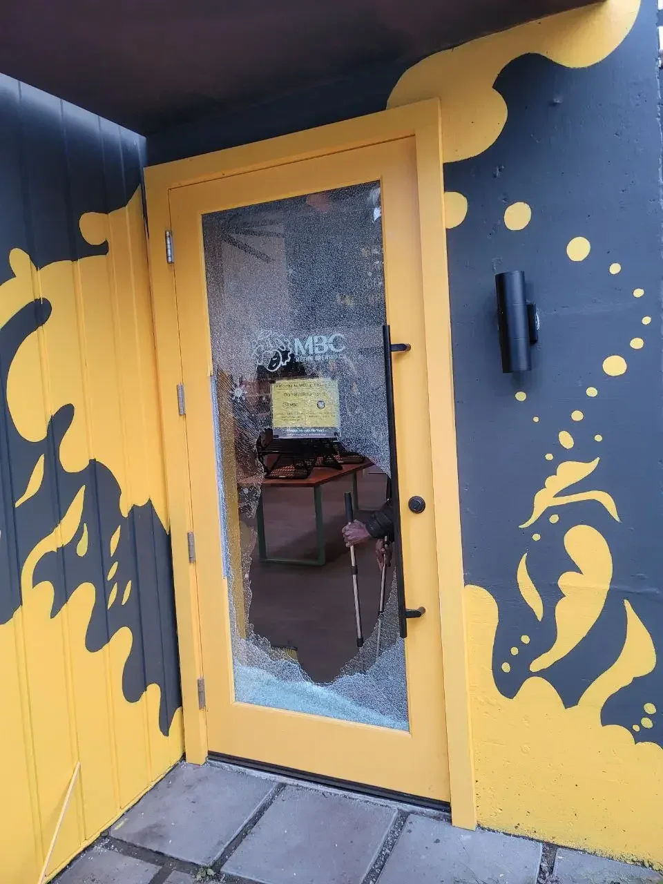 Yellow door with shattered glass around a large opening, against a blue and yellow painted wall.